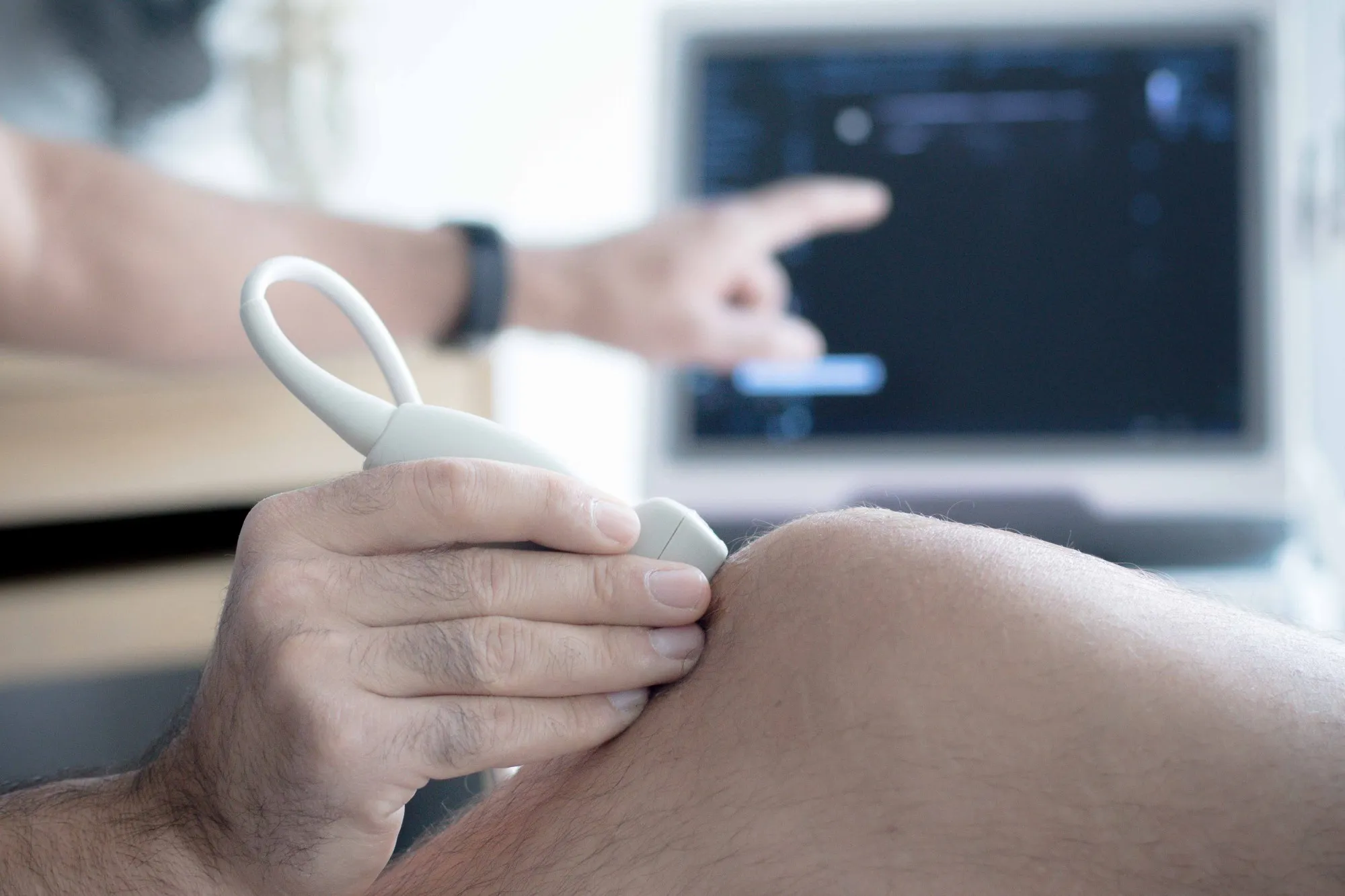 Ultraschalluntersuchung am Knie: Der Schallkopf liegt am Kniegelenk, im Hintergrund zeigt der Arzt auf das Ultraschallbild am Monitor